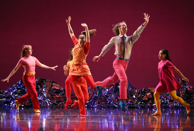 A group of dancers in hues of pink and orange skip and leap